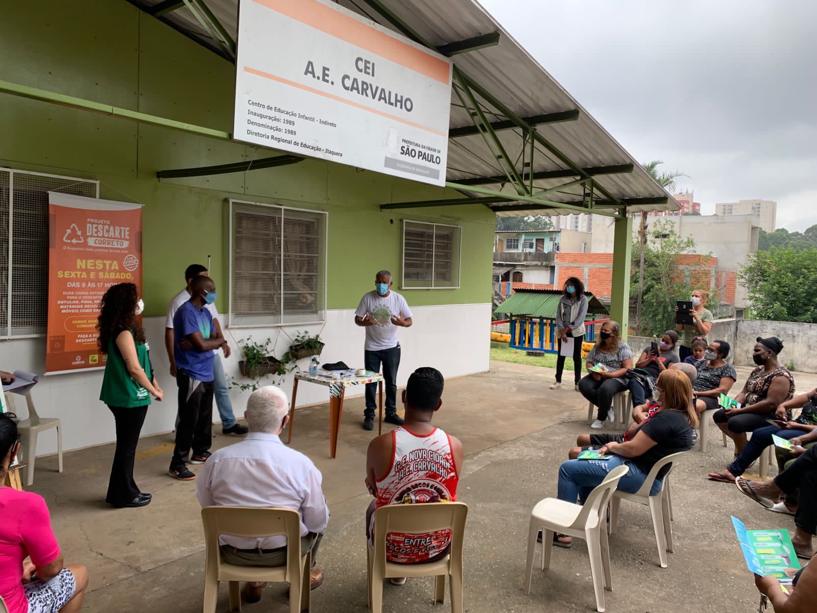 Em um espaço externo coberto do Centro de Educação Infantil (CEI) A.E. Carvalho, em São Paulo, um grupo de pessoas participa de uma palestra sobre descarte correto de resíduos. Um homem de máscara, segurando um objeto, conduz a apresentação enquanto os participantes, sentados em cadeiras plásticas, acompanham atentamente. Ao lado, um banner do "Projeto Descarte Correto" informa sobre horários e instruções para descarte de materiais recicláveis. Algumas pessoas tiram fotos, e o ambiente ao redor inclui edificações residenciais e uma pequena área de lazer infantil.
